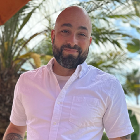 Man with a beard wearing a light pink shirt outdoors with palm trees in the background