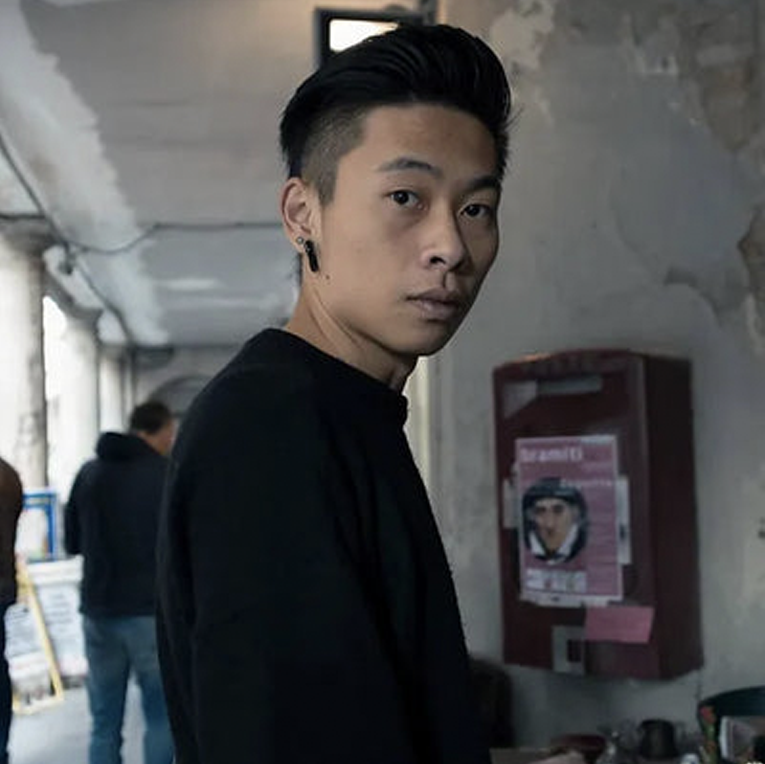 Man in a black shirt standing in an urban setting with a poster on the wall.
