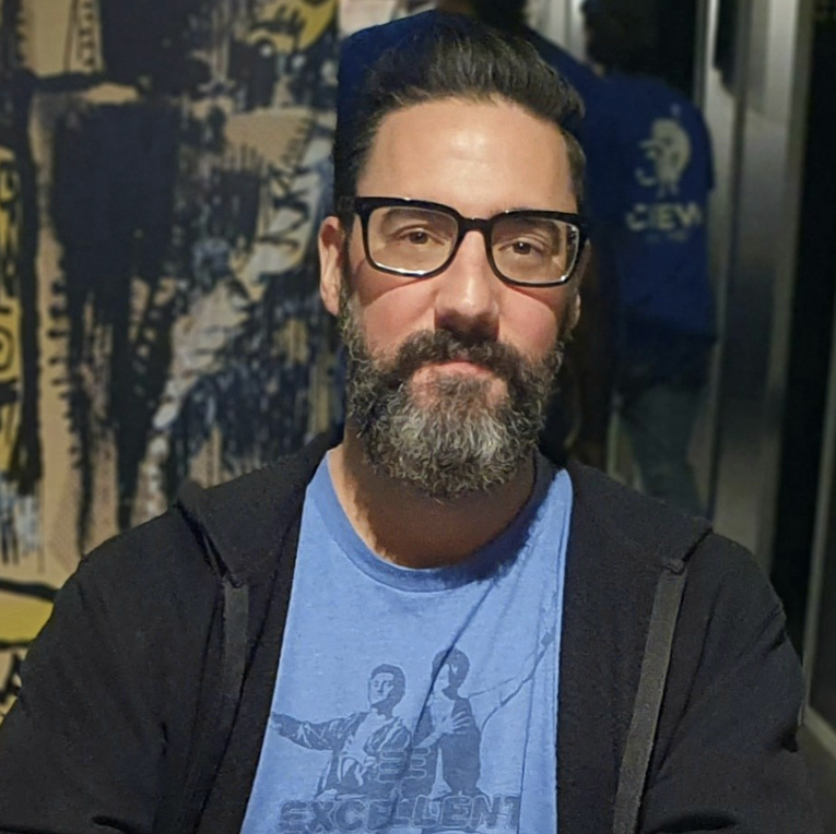 Man wearing glasses and a blue t-shirt with graphics and text, standing in front of a wall with artwork.