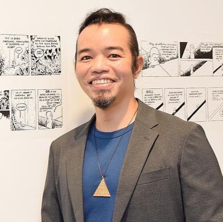 Man in a gray blazer over a blue shirt with comic book panels on the wall behind him