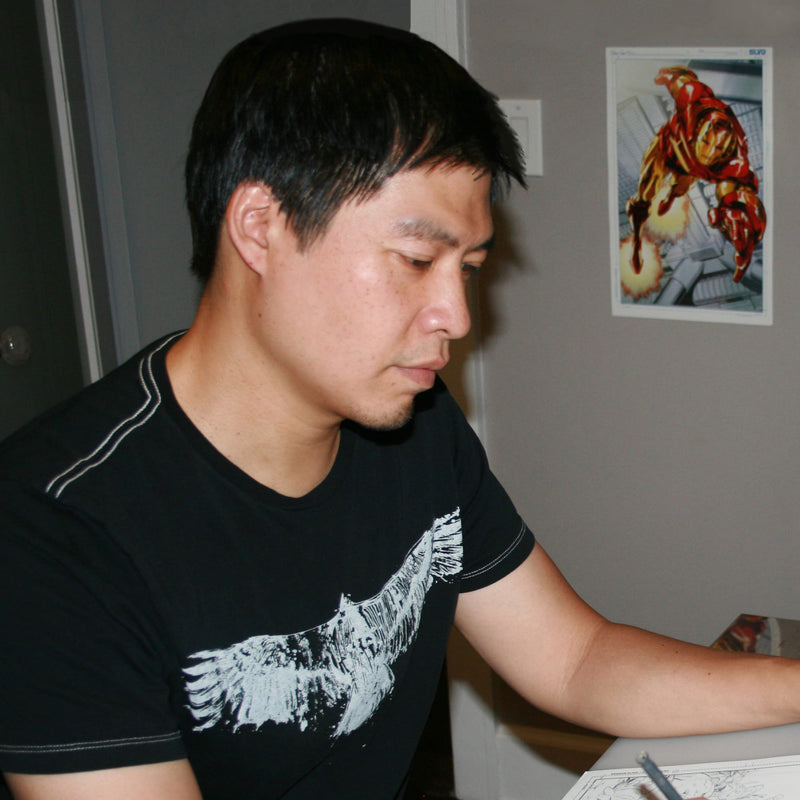 Man wearing a black t-shirt with a white graphic, sitting at a table with a colorful poster on the wall behind him.